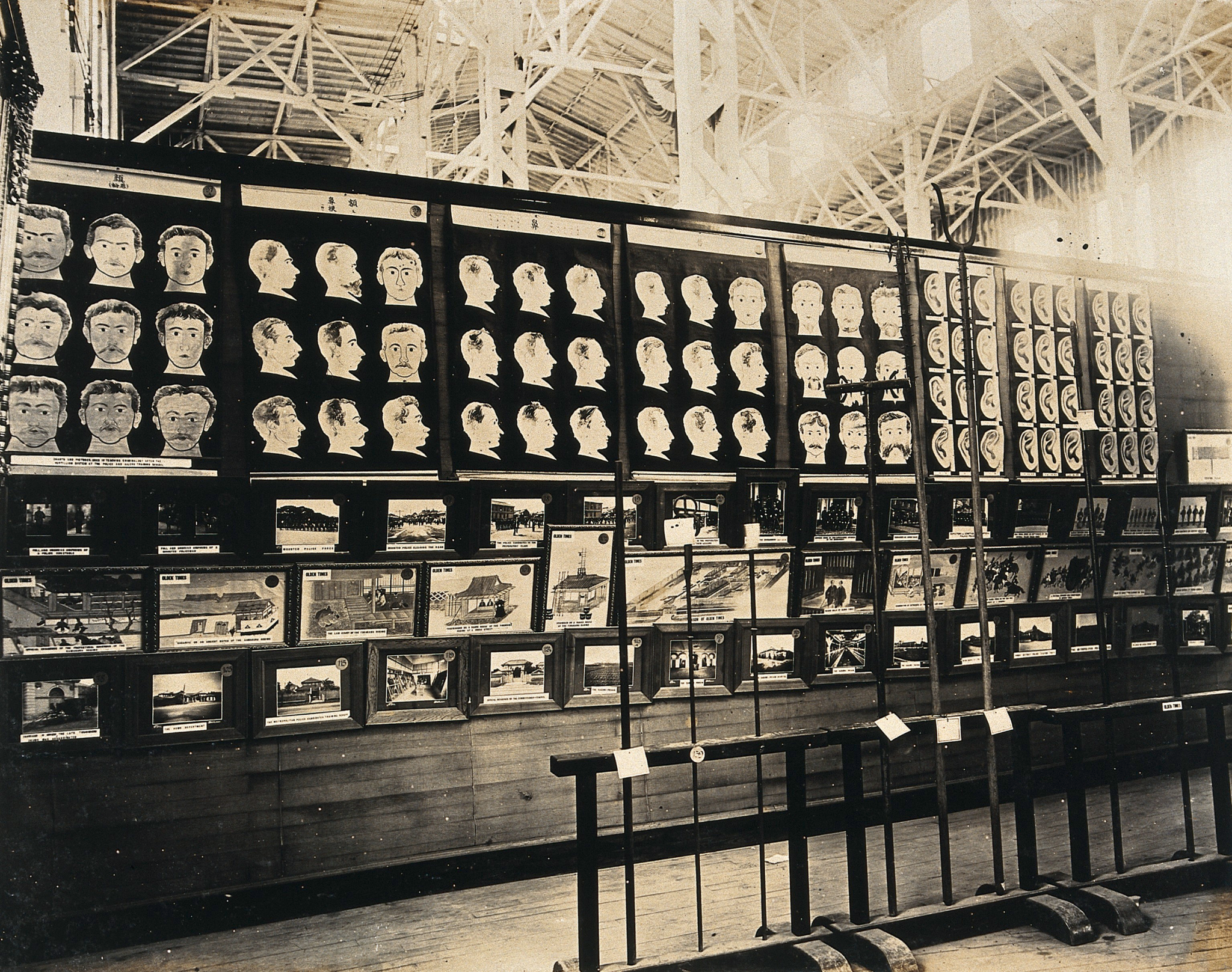 A wall panel containing images of the frontal and profile views of human heads on the top half of the wall, and three rows of photographs of numerous buildings below. The wall and barricades in front of the wall are contained inside a large warehouse-type building.