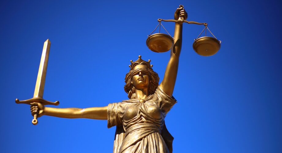 Photograph capturing a gilded statue of Lady Justice, blindfolded and wielding a sword in her outstretched right arm, while delicately holding a balanced scale in her left hand raised above her head.