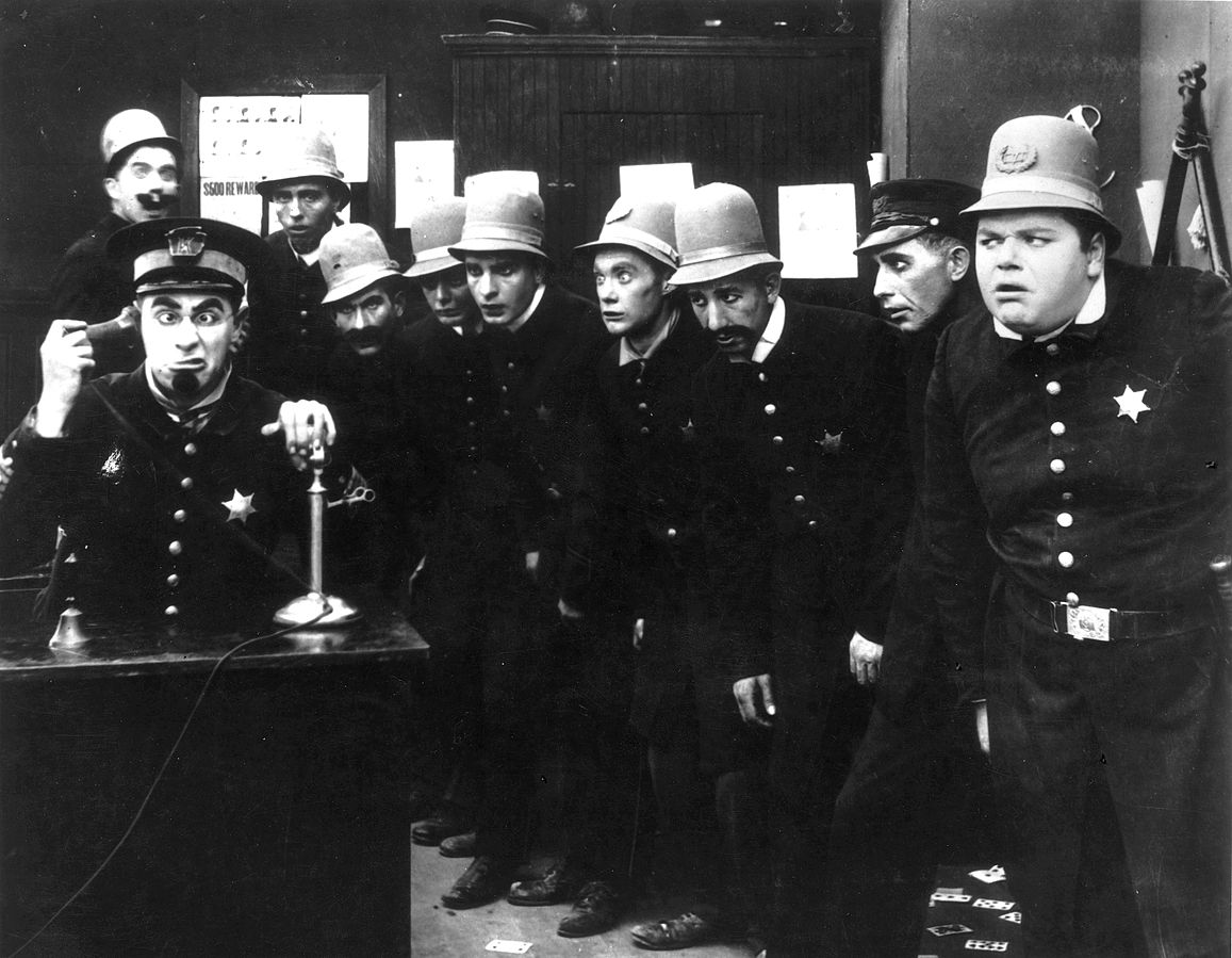 Freeze frame from an early 20th-century silent film depicting a large group of incompetent policemen or Keystone Cops for comedic satire.