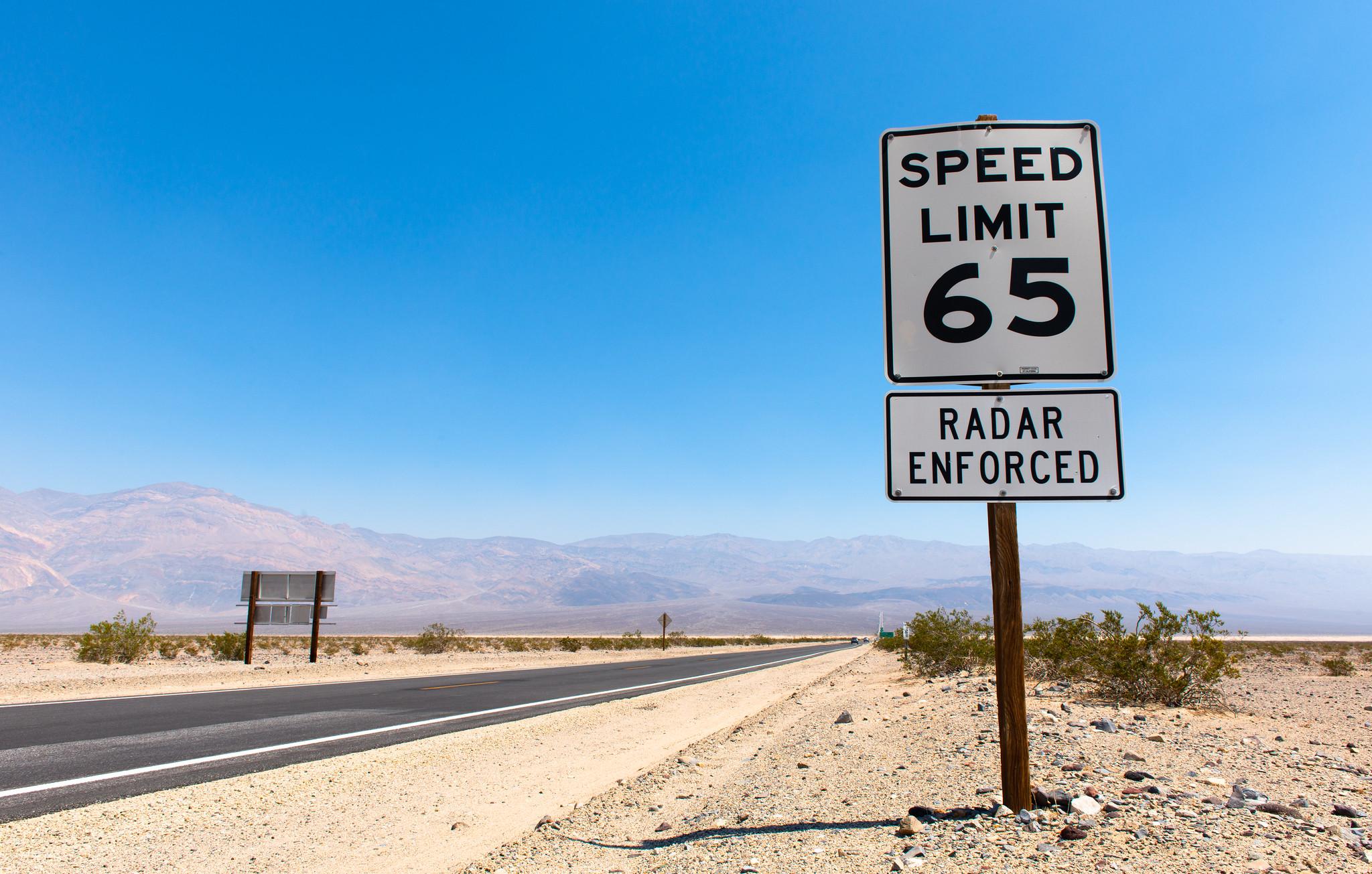 A white speed limit sign with black letters reading “Speed Limit 65” positioned above a smaller sign reading “Radar Enforced”, both signs fastened on a vertical post. A flat, desert-like landscape with low brush in spots on the road sides, with a low mountain range in the background.