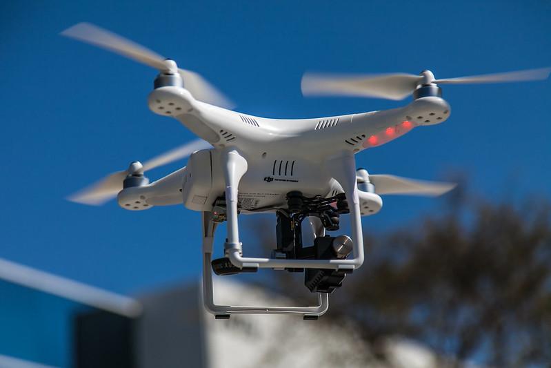 A white unmanned aerial vehicle (UAV), also known as a “drone”, hovers in the air with a building and a tree in the background.