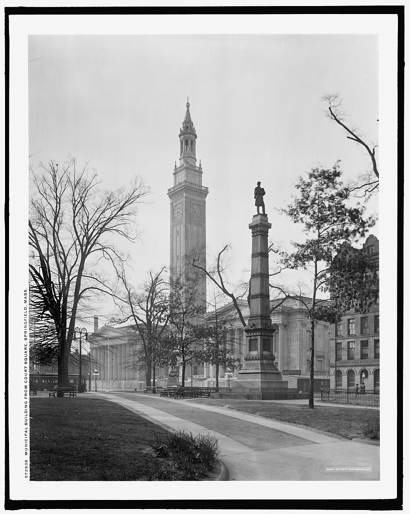 Captivating photograph artfully showcases the historic Municipal Building at Court Square in Springfield, Massachusetts. The stately structure is adorned with Roman columns flanking each side of a lofty clocktower, featuring Gothic architecture, adding to the grandeur of the scene. In the foreground, a towering Civil War memorial statue commanding attention in the foreground, adding to the overall majesty of the scene.