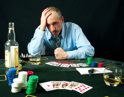 A man in a blue shirt sits at a gambling table. On the table are playing cards, poker chips, a set of car keys, an open liquor bottle which is nearly empty, and two glasses.