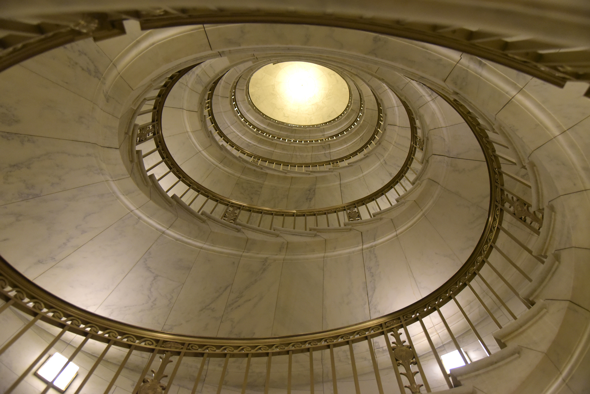 Photograph capturing the breathtaking architecture within the interior of the U.S. Supreme Court Building, featuring a marble staircase adorned with Roman-styled bronze banisters spiraling for several floors toward a glistening golden ceiling in the distance.