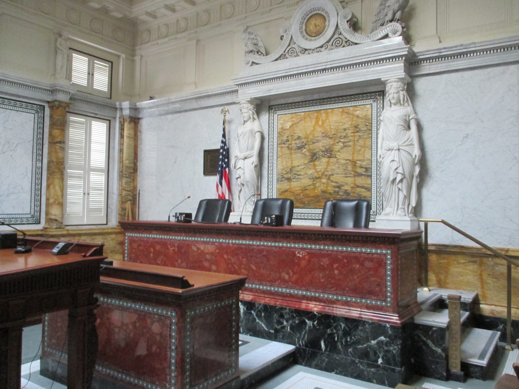 Photograph capturing the interior of a courtroom showcasing notable features, including intricate carvings and two marble statues adorning the wall behind the three-seated judge podium.