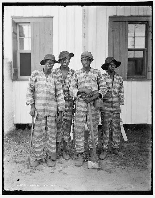 Photograph of four young African Americans chained together at the ankle, clad in dirty, horizontal striped prison uniforms, and holding axes and other labor tools.