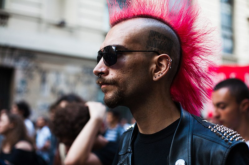 A man with a bright pink Mohawk-style hairstyle, wearing sunglasses and dressed in a metal-studded leather jacket, is standing in a crowd of people. He has a barcode tattooed on his head above his left ear.