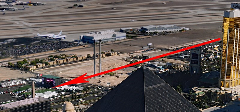 A suburban Las Vegas landscape, with a golden high-rise hotel on the right side of the photo, a black peaked roof in the center foreground, and a red arrow drawn pointing from the hotel to the lower left to a field area. This depicts the line of sight of the shooter from the 32nd floor of the Mandalay Bay Resort and Hotel Complex