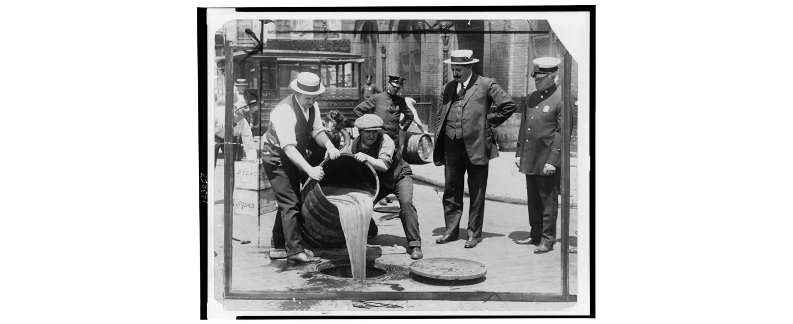 The Deputy Chief of the New York Police Commission stands in the street, monitoring two civilians as they pour a barrel of liquor into the sewer through an open manhole, while two other officers watch nearby.