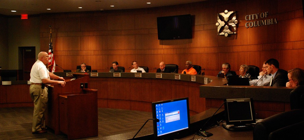 Columbia, MO Police Chief speaks to the Citizens Police Review Board. The Chief stands at a podium facing ten men and women Board members seated behind a counter.