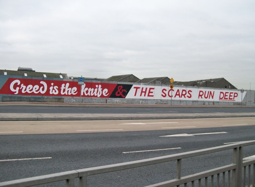 Image showing graffiti along the walls lining a freeway that reads, “Greed is the knife & the scars run deep.”