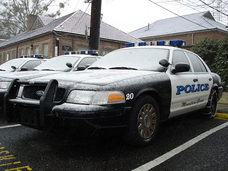 Three black and white police cars are backed in to respective parking stalls next to each other. The car in the foreground has the number 20 on its front fender near the headlight. Two beige-colored office buildings and a power pole are seen behind the cars