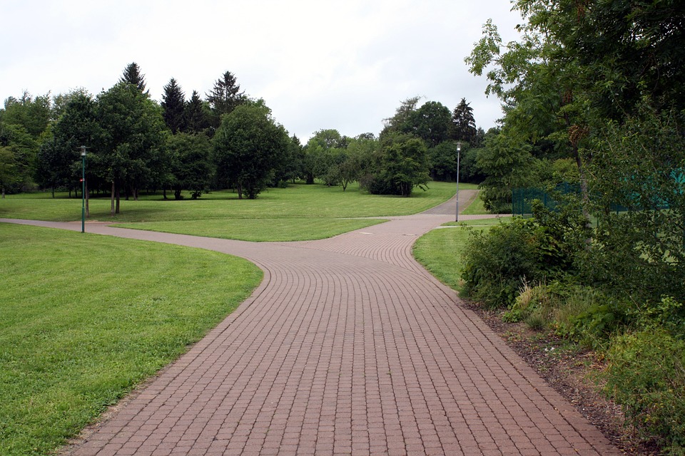 A wide brick pathway in a park leads to a path to the left, and one to the right. Trees and green grass are on the sides of the path and background. A light standard is located on the right side of the right-hand path.