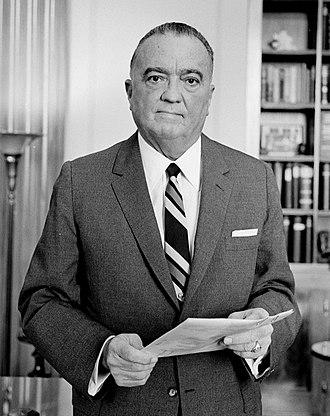 Photograph of J. Edgar Hoover standing, appearing stoic, dressed in a professional suit and tie, holding a document in his hands, with rows of shelved books in the background.