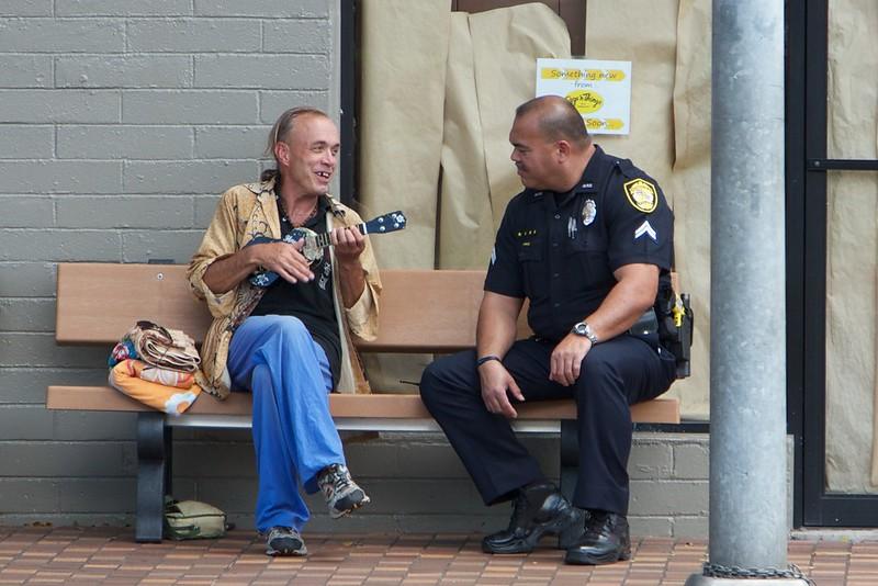 A man, seated on a bench, is playing a ukelele speaks with a police officer seated on his left.