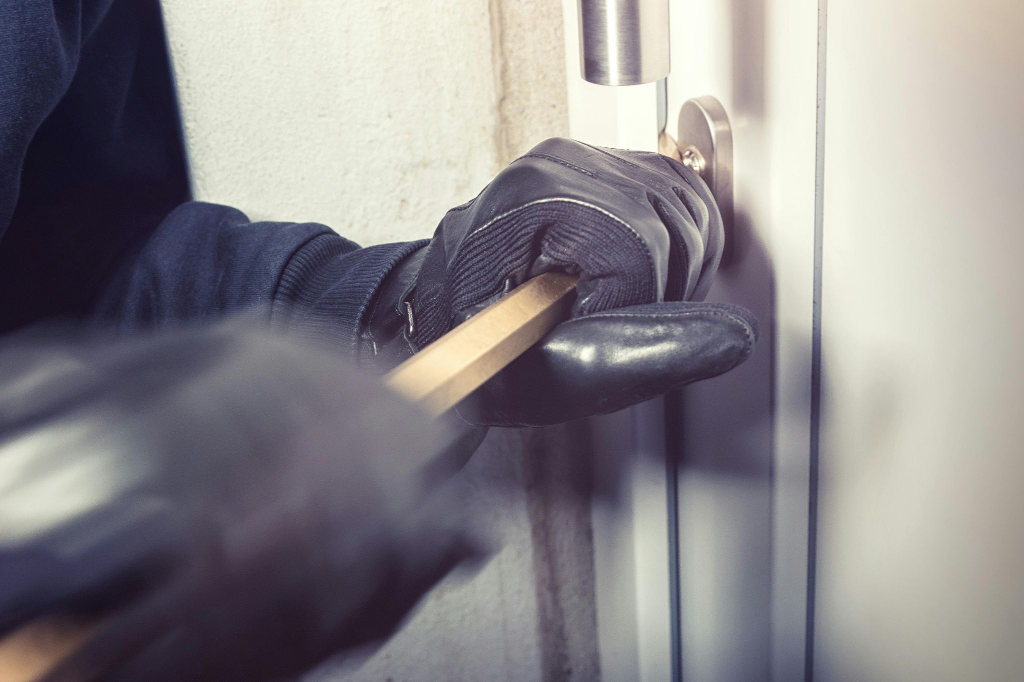 Close-up of black-gloved hands using a pry bar to forcibly open a locked exterior door.