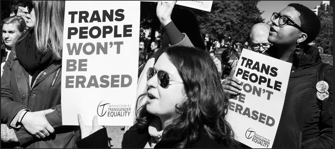 A large group of people who represent the transgender population are demonstrating in Washington D.C. Several members are holding white posters that read “Trans People Won’t Be Erased.” Under these words on the lower right hand corner of each poster is a logo and the words “National Center for Transgender Equality.”