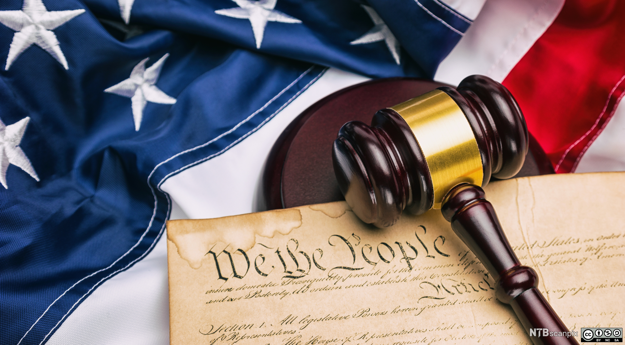 Photograph of a gavel, representing justice, carefully placed across the U.S. Constitution, resting on a backdrop of the majestic American flag.