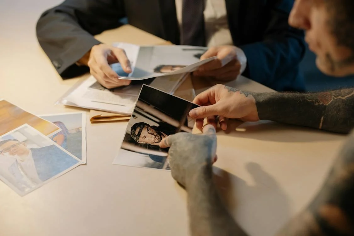 Two people are sitting at a table looking at photographs of potential suspects in a crime. The man in the foreground is pointing toward a suspect in the photo he is holding. He has a dark sleeve tattoo on his right arm. The man seated in the rear of the photo is holding more photos, and he is wearing a dark coat and tie.