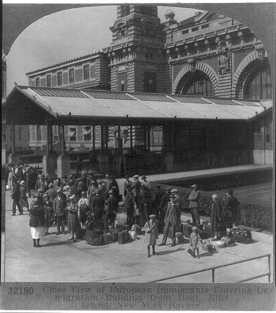 Photograph of dozens of European immigrant families with their luggage, waiting outside the Ellis Island Immigration Station on a sunny July day.