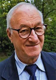 Albert Bandura, a middle-aged male, is wearing a dark suit jacket, light blue shirt, dark tie, and glasses.