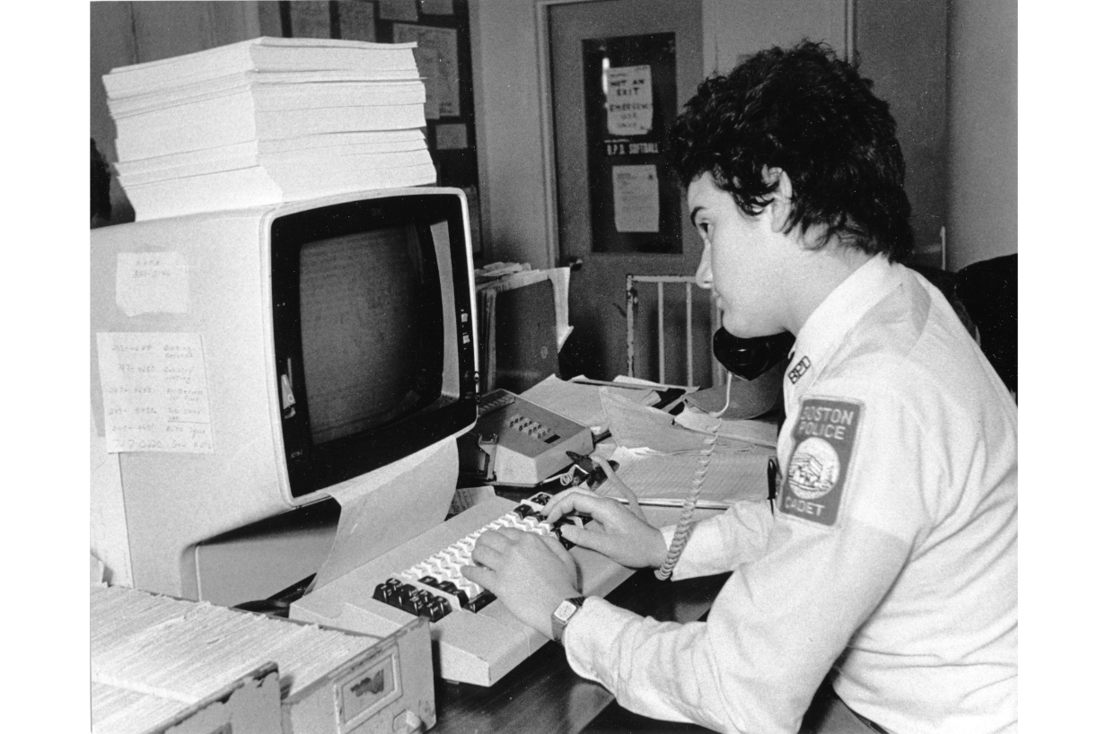 Photograph of a Boston Police Department female cadet learning new computer technologies at a document-laden desk.