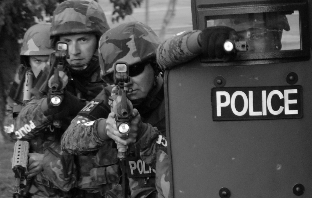 Three officers in tactical uniforms and holding rifles stand alongside a fourth officer, who is in front wearing an armored shield.