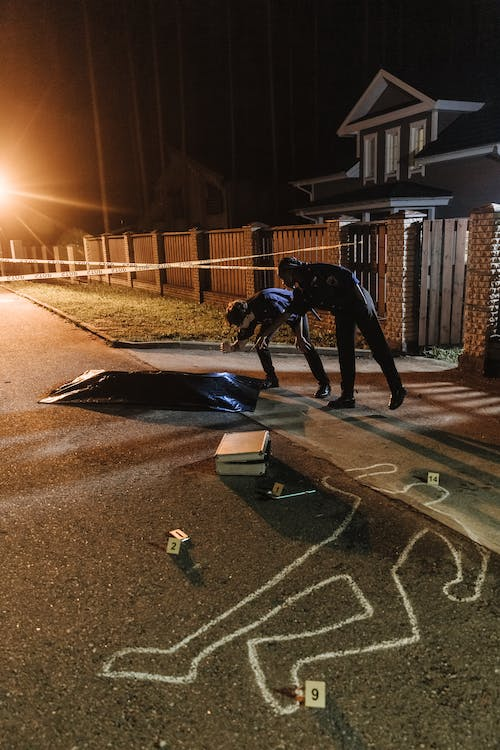 Two officers work the scene of an apparent homicide. A chalk sketch appears on the street up onto the sidewalk, with four plastic evidence tents at specific sites of evidence. A partially-opened briefcase is on the street, lying between a black body bag and the chalk sketch. Crime scene stape is stretched and fastened to some brick entrance gates.