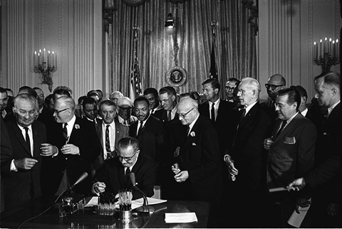 President Lyndon B. Johnson, pen in hand, signs the 1964 Civil Rights Act as Martin Luther King, Jr. and other political figures eagerly watch.