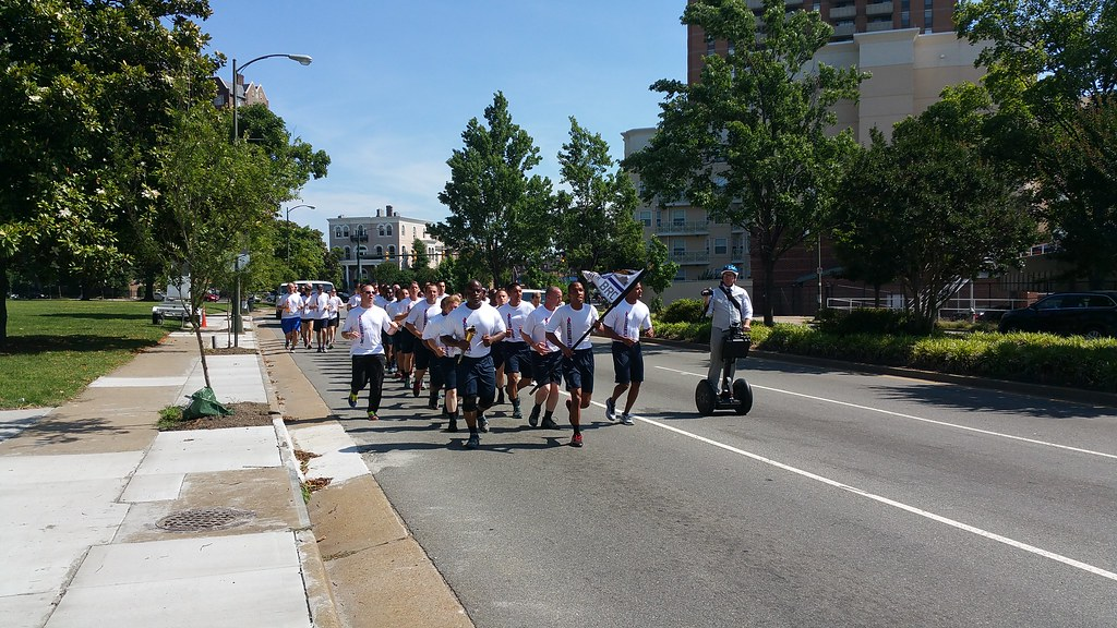 A group of about 20 police recruits dressed in white tee shirts and dark blue shorts run down a city street. The group leader carries a flag, and a man riding to the side on a segway takes photographs.