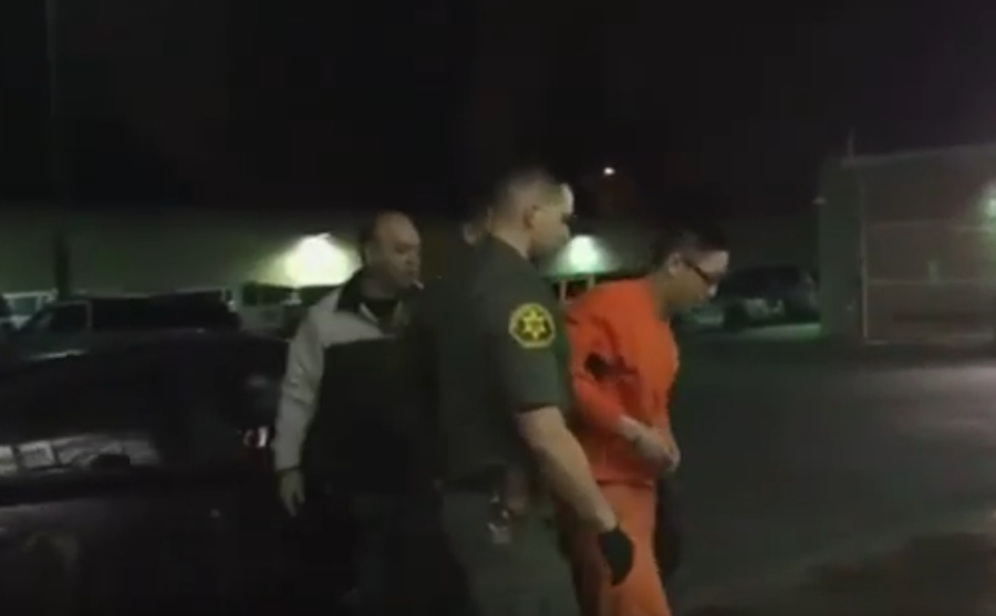 Image of a parolee in handcuffs wearing an orange jumpsuit, escorted by corrections officers.