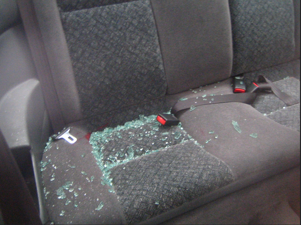 A vehicle rear seat, gray in color, has many fragments of broken glass on it, indicative of a break-in from outside the vehicle. Loose seat belts are also visible.