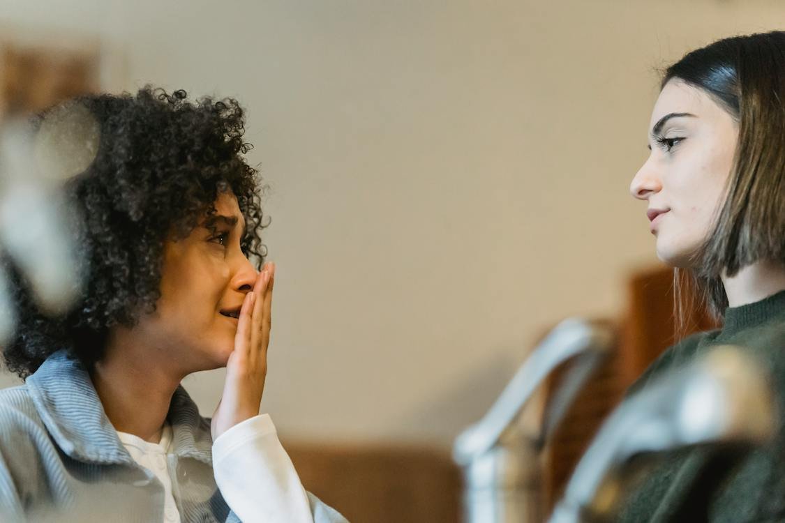 Two women are seated in a room. The black woman on the left is holding her left hand vertically in an effort to cover her mouth, and appears to be in grief. The Caucasian woman on the right is looking at the other woman and attempting to validate her feelings.