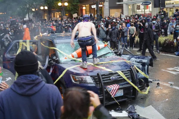 A shirtless man, facing away, stands on the hood of a blue vandalized police vehicle, which has yellow tape criss-crossing it, an American flag in the grille, and traffic cones through the windshield. A large crowd is in the background.