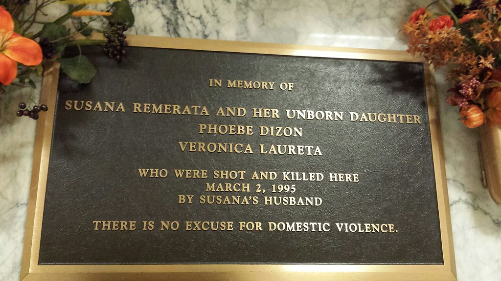 A rectangular bronze plaque in memory of Susana Remerata, her unborn daughter, and two other women, is displayed on a white marble background. Flowers adorn both upper corners.