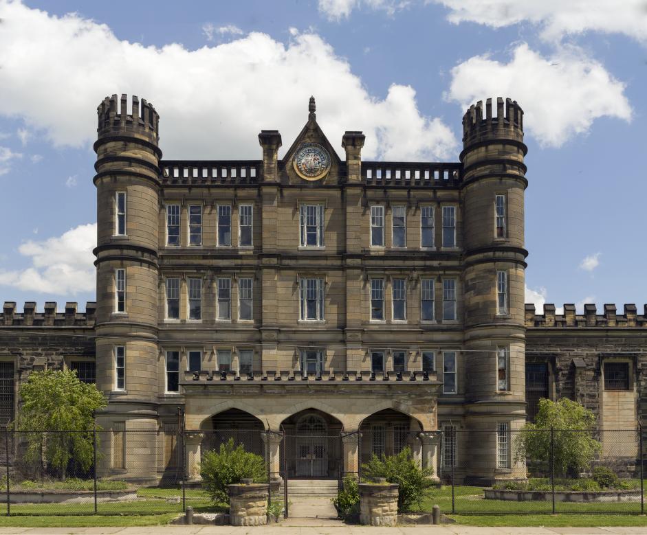 Photograph of the exterior of the West Virginia State Penitentiary featuring a grand, Gothic-style building with pointed arches, intricate stone carvings, and tall spires, surrounded by a high chain-linked fence.