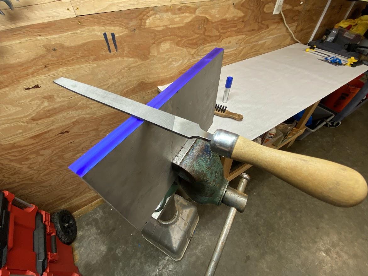 A plate of aluminum stock clamped in a vise with layout fluid applied to an edge with a file resting on the edge.