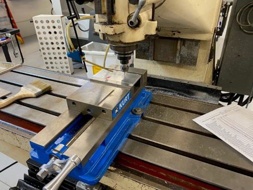 A CNC bed mill has a vise bolted to the worktable with a 3 by 6-inch piece of aluminum secured in its jaws. An engraving tool contacts the stock surface and engraves letters onto the surface of the metal.