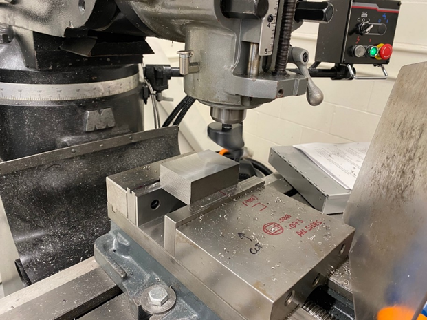 A piece of steel is held in a machine vise, and a fly cutter tool is rotated and advanced across the part to create a flat and smooth surface.