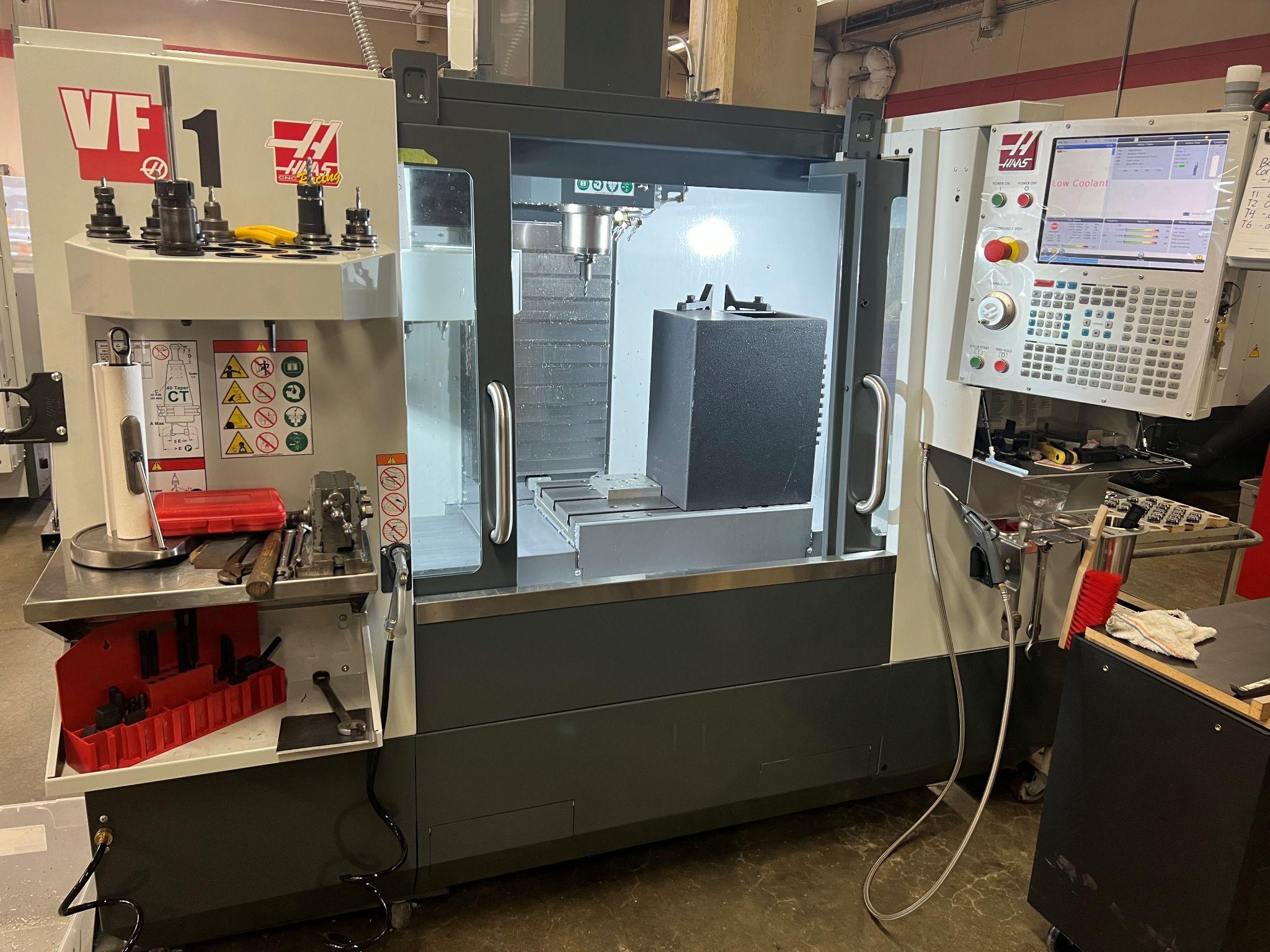 A CNC Mill. The light is on inside the machine where the parts are made. There is a big block of material sitting on the right side of the table. The controls to run it are on the right side, and a shelf of tools and clamping hardware is on the left.