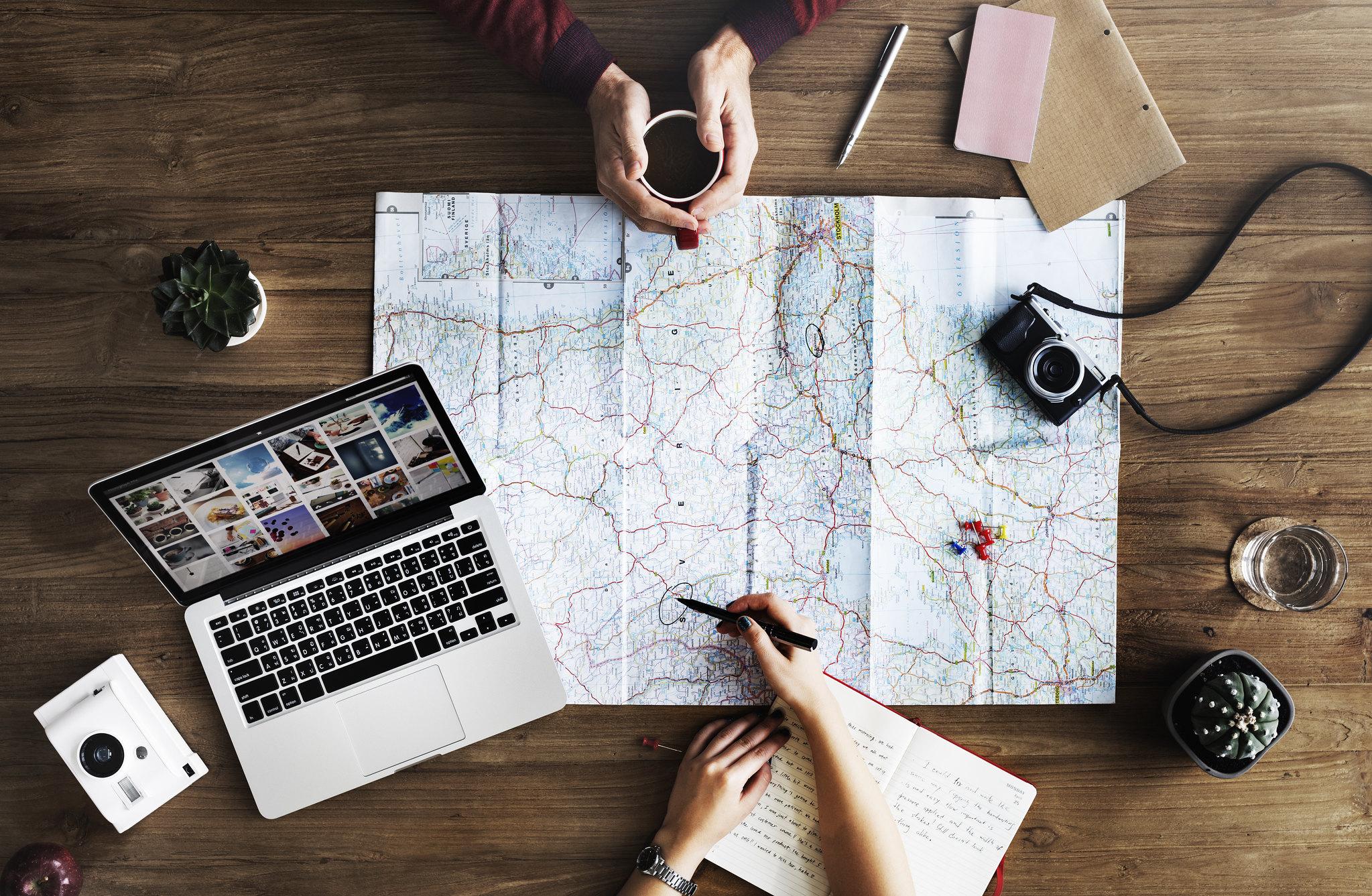 Table with map, laptop, notepad, cameras, plants, cup of coffee, and water. Two pairs of hands, one holding a pen, implying that they are looking at a map in preparation for a trip.