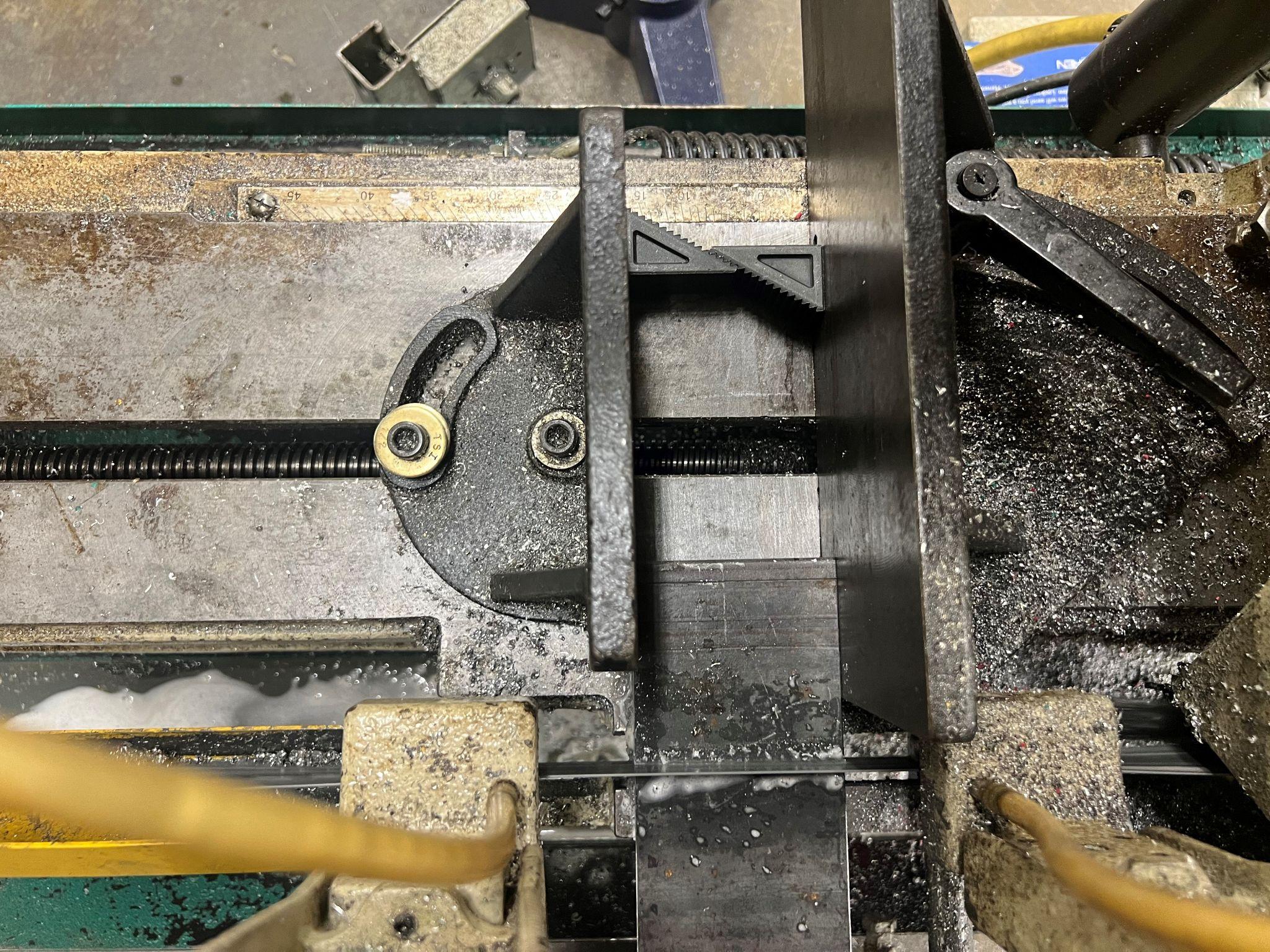 Horizontal bandsaw cutting metal with coolant running across the part. This is looking directly down at the part. The part is at the bottom, and the step blocks are at the top. The pivot point of the vise can be seen.