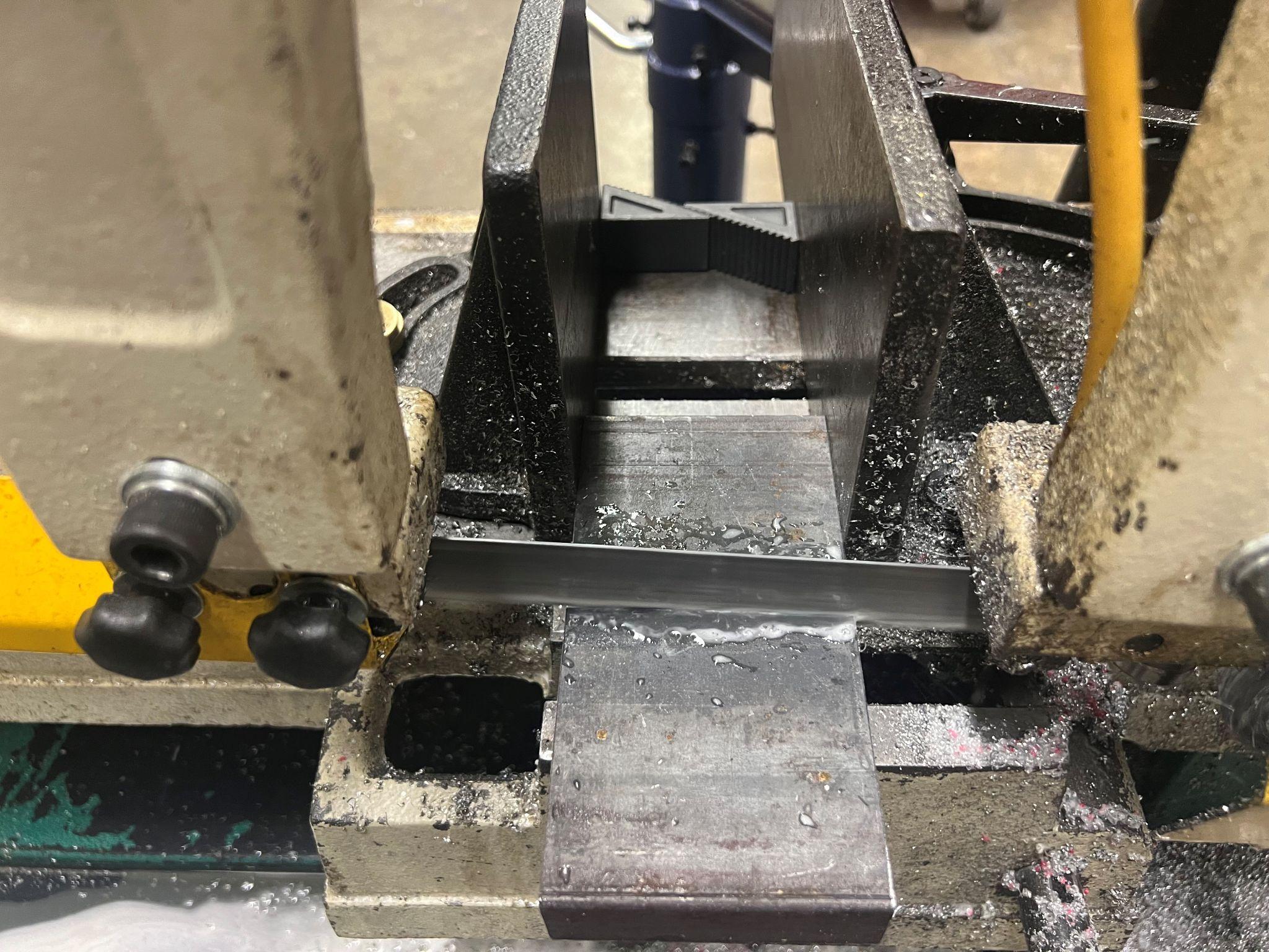 Horizontal bandsaw cutting metal with coolant running across the part. The guides are positioned close to the part to support the blade. Step blocks are on the opposite side of the material being cut in the vise.