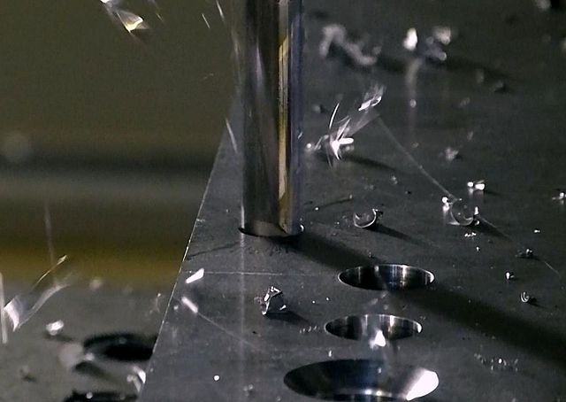 A drill is cutting into a metal plate. Three holes made by the drill precede the one being drilled. There are metal chips falling around the work surface.