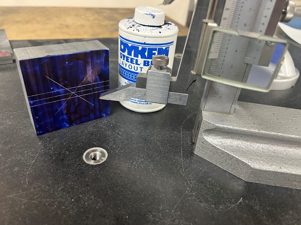 From left to right, a part with scribe lines indicating the center of the part, and the width of the V groove. A bottle of Dykem, blue layout fluid, and Vernier scale height gauge.