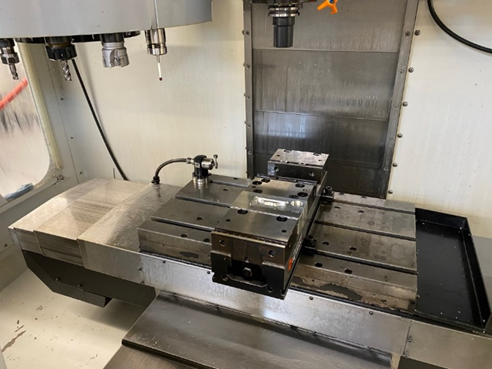 A two position vise rests on the worktable of a CNC mill. Tools are visible in the carousel above the worktable.