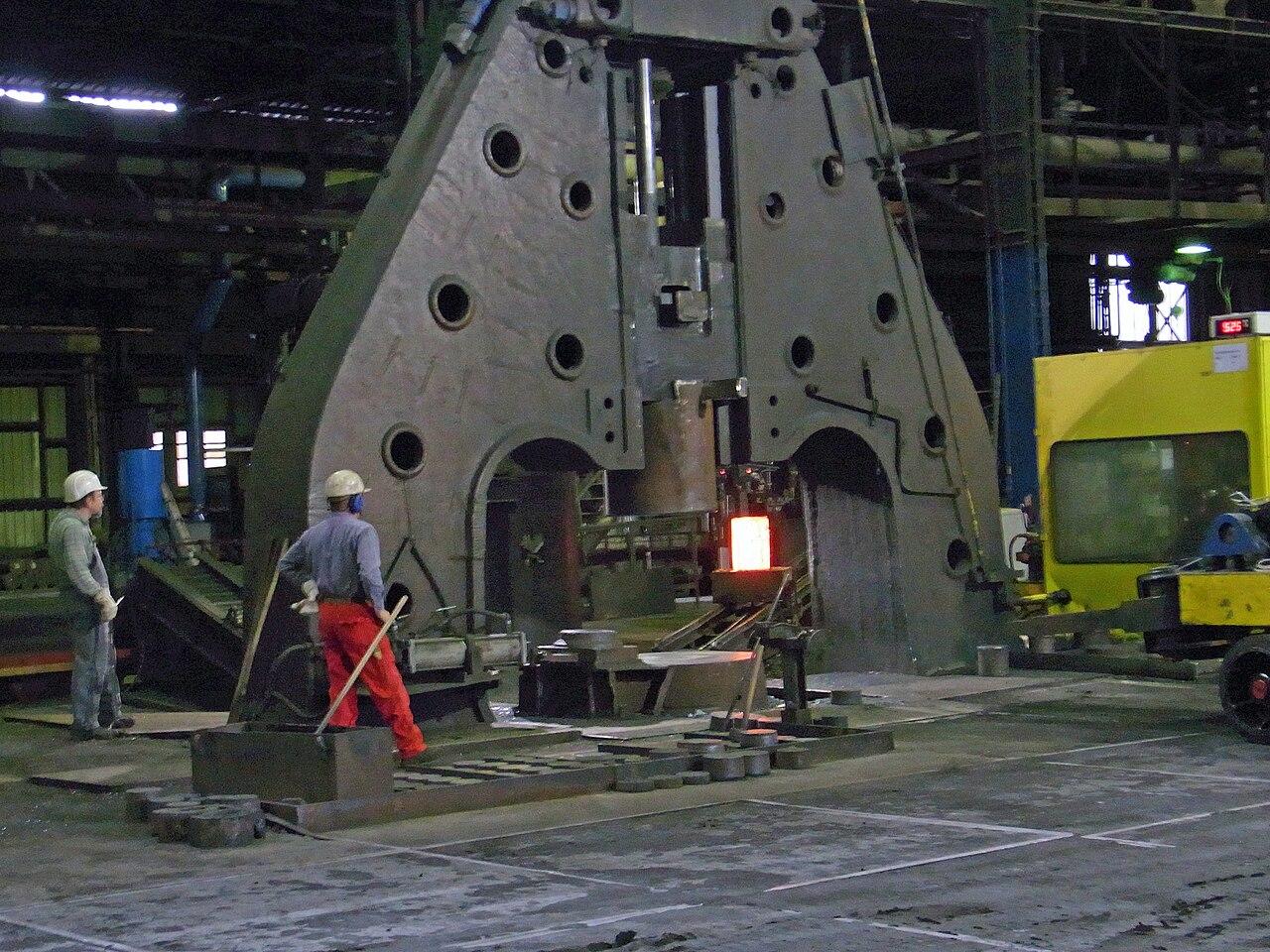 A hot metal ingot is being loaded into a hammer forge.