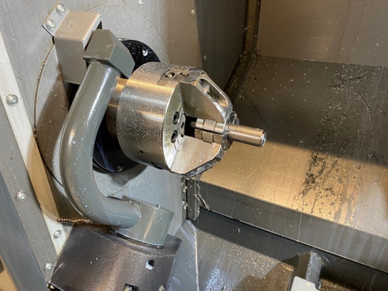 A short piece of steel is held in a three-jaw chuck inside a CNC lathe enclosure. Coolant droplets cling to all the surfaces,and metal chips are resting on the flat surfaces.