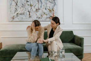 Two women seated on a couch in a white office. The woman on the left is holding her hand to her head and appears distraught. The woman on the right has her right hand on the other woman’s right shoulder, and is holding her left hand so as to comfort her.