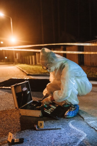 A crime scene investigator, clothed in a Tyvek suit and shoe coverings, squats near an open briefcase containing fingerprinting materials. In the foreground are two evidence tents, with one associated with a tire iron. The pavement also has chalk marks depicting the final resting point of a person who evidently was a victim of assault.